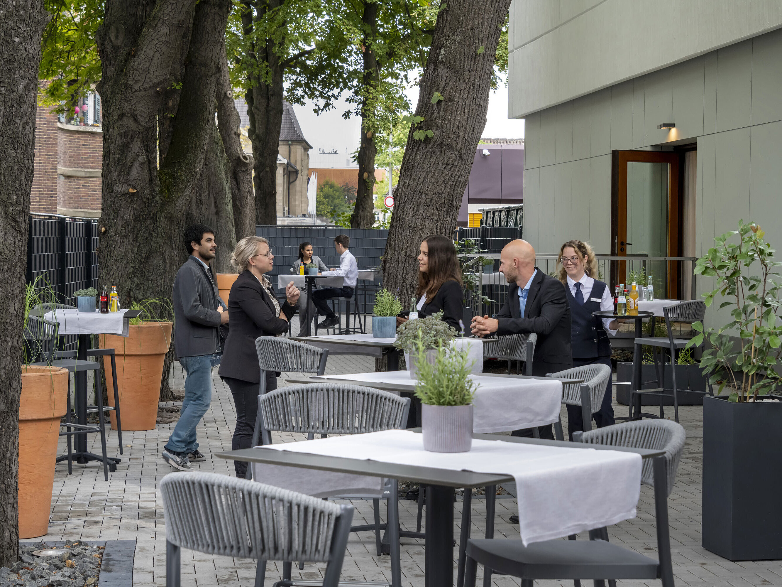 Moderne Terrasse im Maritim Hotel Darmstadt mit Gästen, die entspannen, Bäumen im Hintergrund und stilvollen grauen Möbeln.